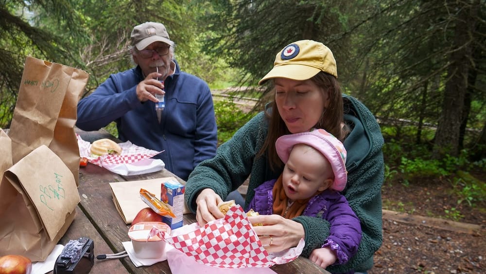 Audrey Bergner sits with Aurelia Jeffery at a forest picnic table near Moraine Lake in Banff National Park, Alberta, Canada, while Daniel Bergner eats lunch beside them.