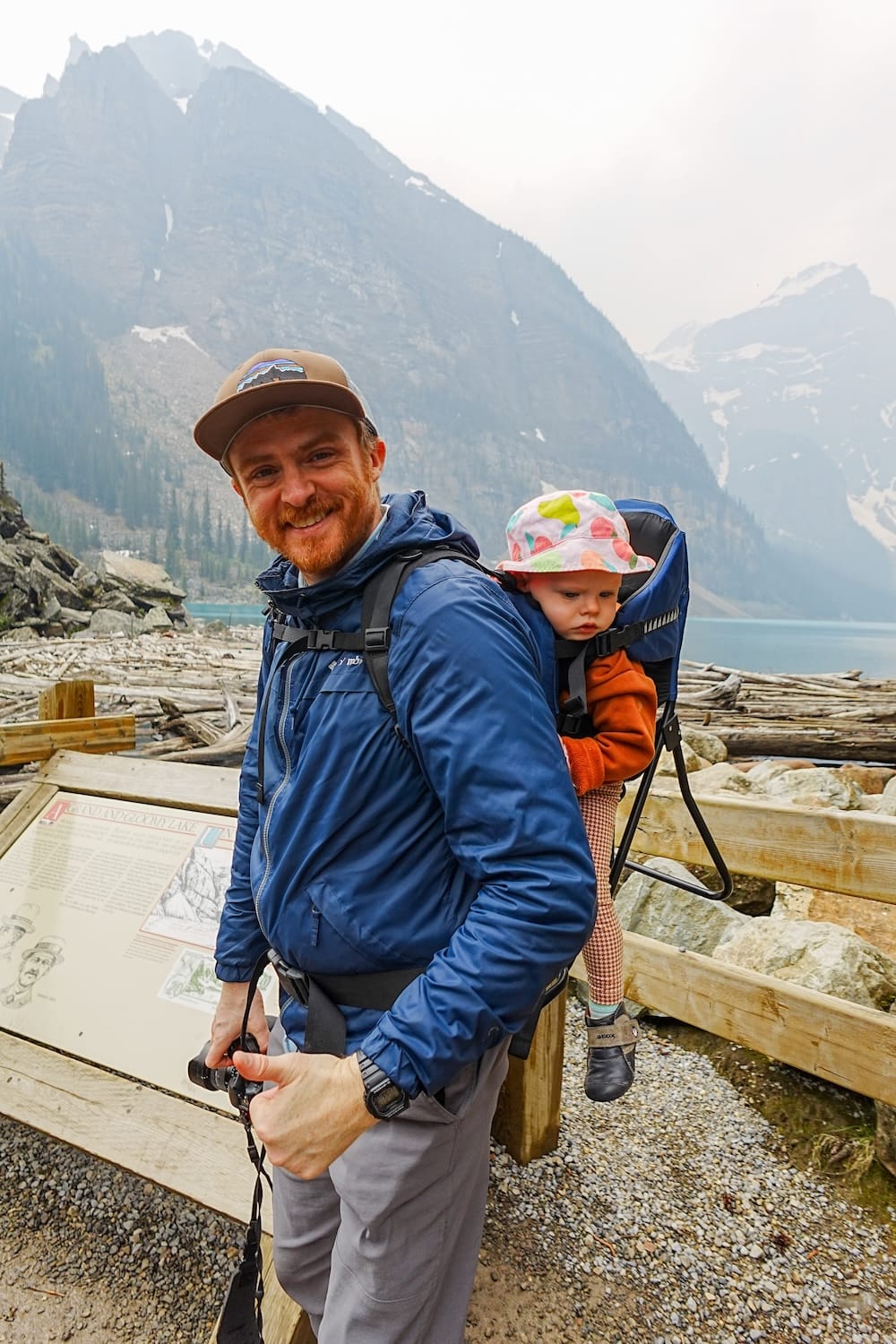 Samuel Jeffery lleva a Aurelia Jeffery en una mochila portabebé junto a Moraine Lake, en Banff National Park, Alberta, Canadá, con costa rocosa, agua turquesa y montañas empinadas detrás.
