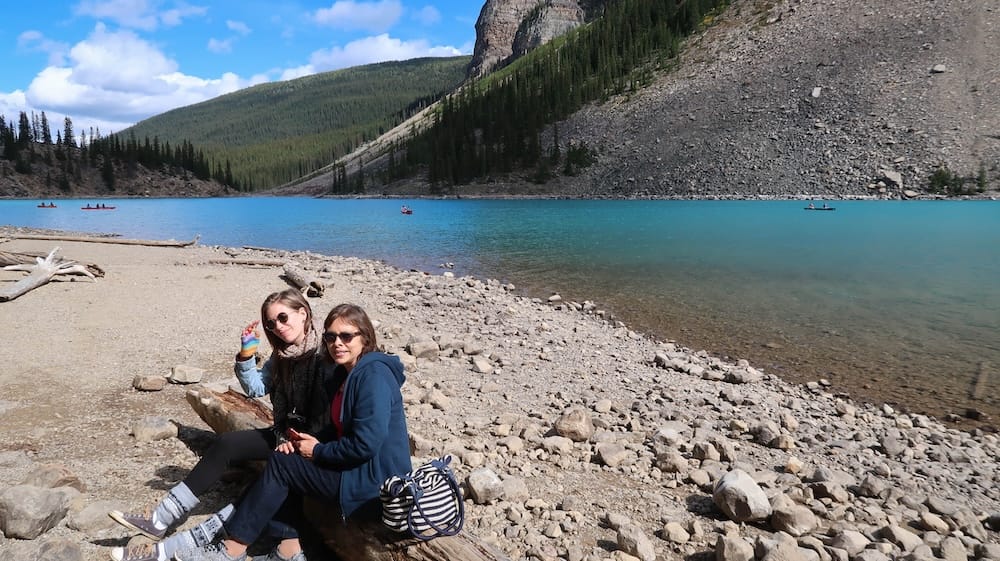 Audrey Bergner sits with her mother on the rocky shoreline of Moraine Lake in Banff National Park, Alberta, Canada, with turquoise water, distant canoes, forest and mountain slopes behind them.