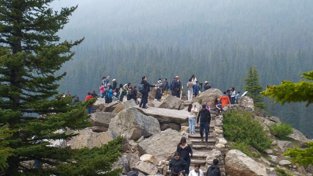 Visitors crowd the rocky Rockpile viewpoint at Moraine Lake in Banff National Park, Alberta, Canada, standing on boulders and stone steps with dense evergreen forest behind them.