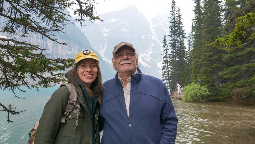 Audrey Bergner and Daniel Bergner at Moraine Lake in Banff National Park Alberta Canada with mountain scenery Audrey Bergner and Daniel Bergner stand beside Moraine Lake in Banff National Park, Alberta, Canada, with turquoise water, evergreen trees and hazy mountain peaks visible behind them.