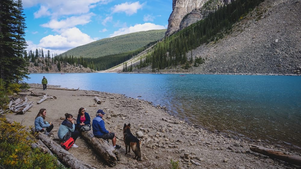 Audrey Bergner, Family and Togo Resting at Moraine Lake, Banff National Park, Alberta, Canada Audrey Bergner rests with family and Togo, their dog, on the rocky shoreline of Moraine Lake in Banff National Park, Alberta, Canada, with turquoise water, forested slopes, and steep mountain walls beyond.