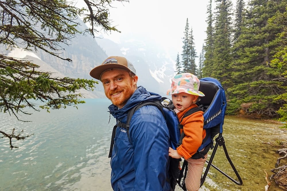 Samuel Jeffery stands beside the shoreline at Moraine Lake in Banff National Park, Alberta, Canada, carrying Aurelia Jeffery in a child backpack with turquoise water, evergreen trees and hazy mountain slopes behind them.