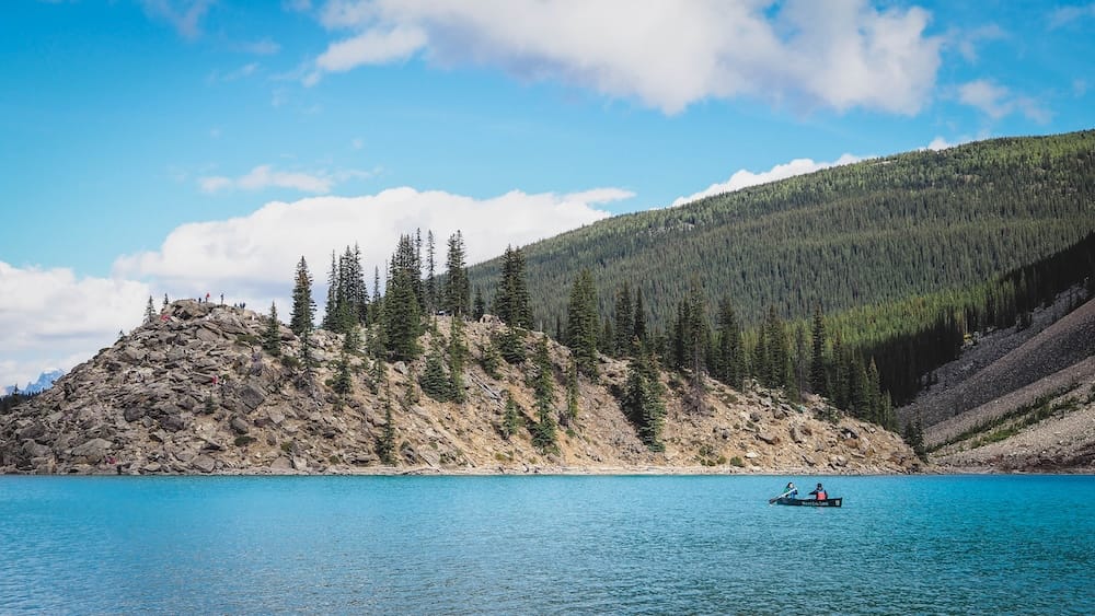 Moraine Lake Turquoise Water and Canoe Below the Rockpile in Banff National Park, Alberta, Canada A canoe crosses the vivid turquoise water of Moraine Lake in Banff National Park, Alberta, Canada, with the rocky Rockpile shoreline, evergreen forest and mountain slopes behind.