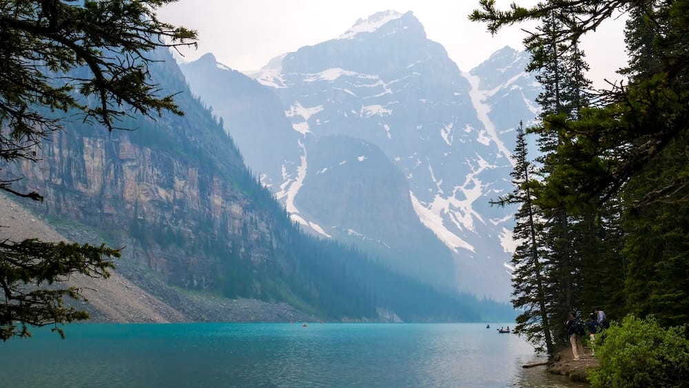 Scenic view across turquoise Moraine Lake in Banff National Park, Alberta, Canada, framed by conifer trees with steep rocky mountains and lingering snow rising behind the water.