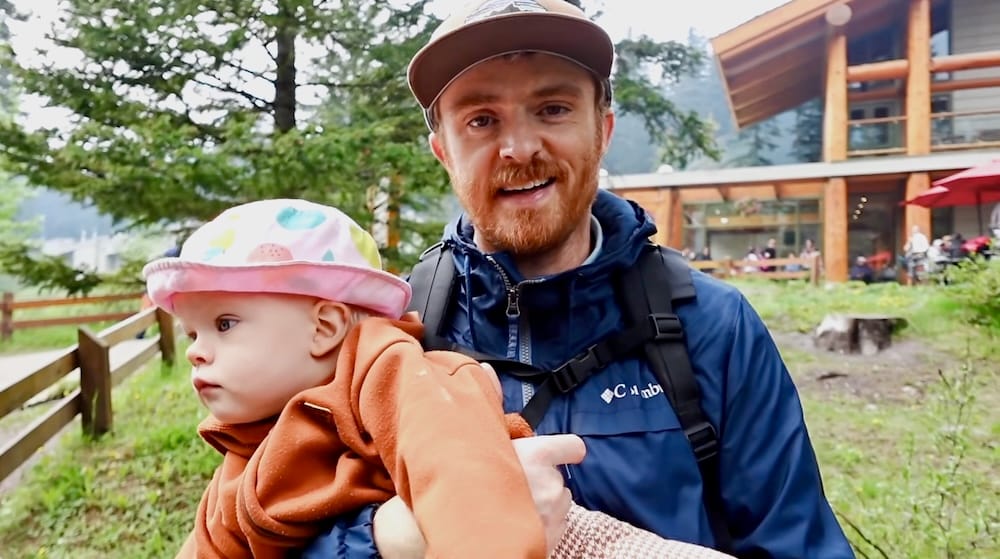 Samuel Jeffery holds Aurelia Jeffery outside Moraine Lake Lodge in Banff National Park, Alberta, Canada, with the timber lodge building, patio area and mountain scenery behind them.