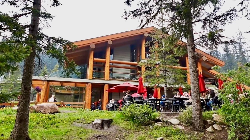 Visitors sit at outdoor tables under red umbrellas on the patio of Moraine Lake Lodge in Banff National Park, Alberta, Canada, surrounded by pine trees and log construction.