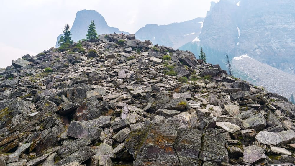 The rocky summit area of the Moraine Lake Rockpile in Banff National Park, Alberta, Canada, rises above a field of broken stone with steep mountain walls and scattered conifer trees behind it.