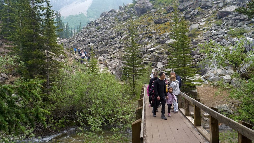 Visitors cross a wooden bridge on the Rockpile Trail at Moraine Lake in Banff National Park, Alberta, Canada, with conifer forest, rocky slopes and more hikers ahead.