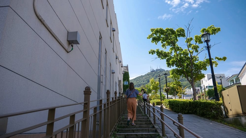 Audrey Bergner climbs a stepped hillside path in Motomachi, Hakodate, Hokkaido, Japan, with railings, street lamps, leafy trees and Mt. Hakodate rising in the background under a bright blue sky.