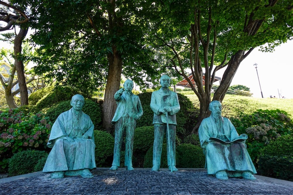 Bronze statues of the Four Heavenly Kings of Hakodate stand beneath leafy trees in Motomachi Park, Hakodate, Hokkaido, Japan, surrounded by landscaped greenery and historic park grounds.