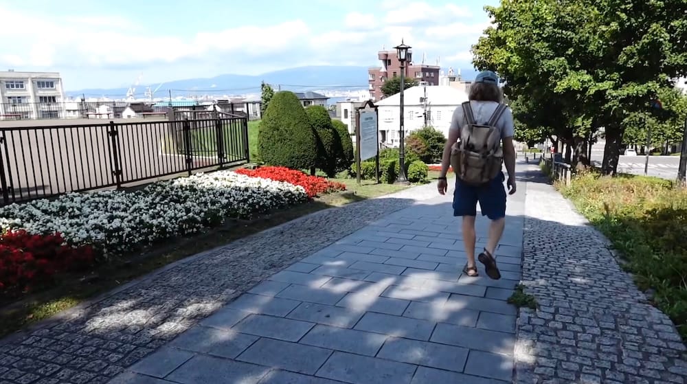 Samuel Jeffery walks away from the camera along a stone-paved path in Motomachi Park in Hakodate, Hokkaido, Japan, past red and white flower beds, trees and a lamp post, with the harbour and mountains beyond.