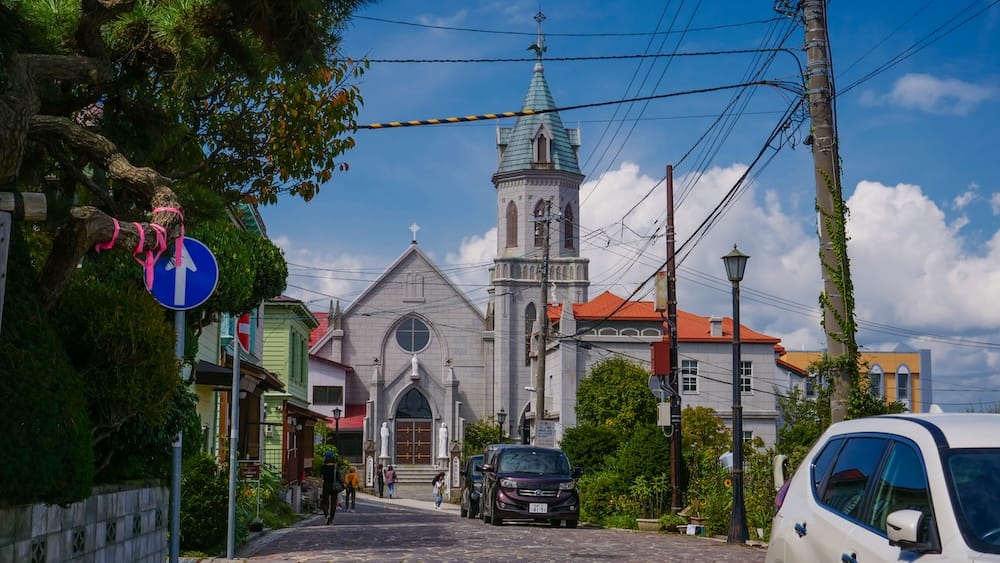 The grey stone Gothic facade and green-spired bell tower of Motomachi Roman Catholic Church in Hakodate, Hokkaido, Japan, seen from a cobbled Motomachi street under a blue sky with scattered clouds.