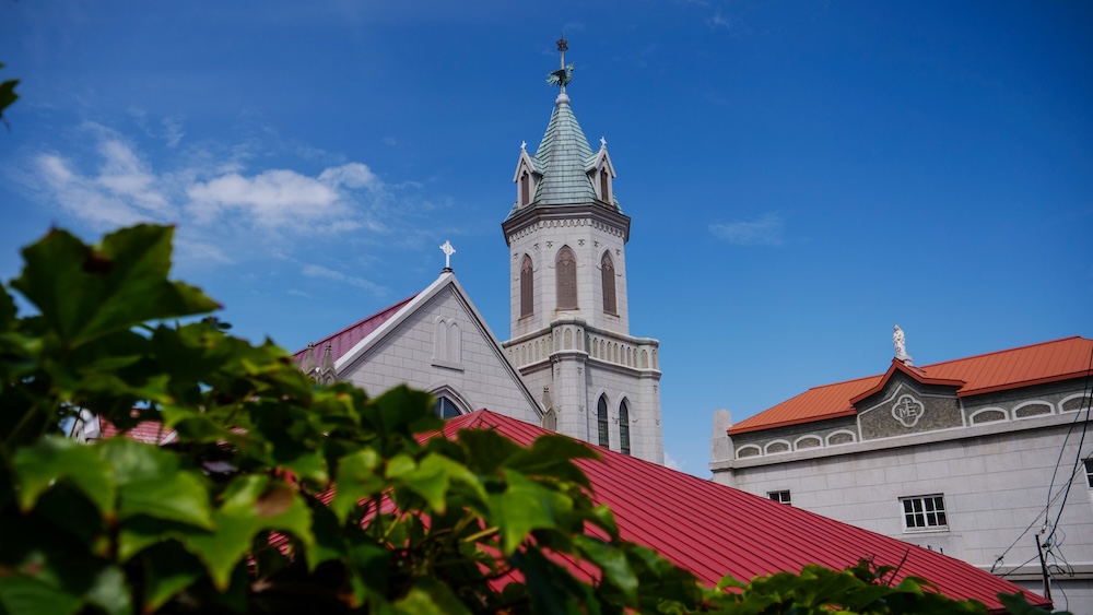 Motomachi Roman Catholic Church in Hakodate, Hokkaido, Japan, with its tall Gothic-style bell tower, green copper spire and white stone exterior rising above red rooftops and leafy greenery against a blue sky.