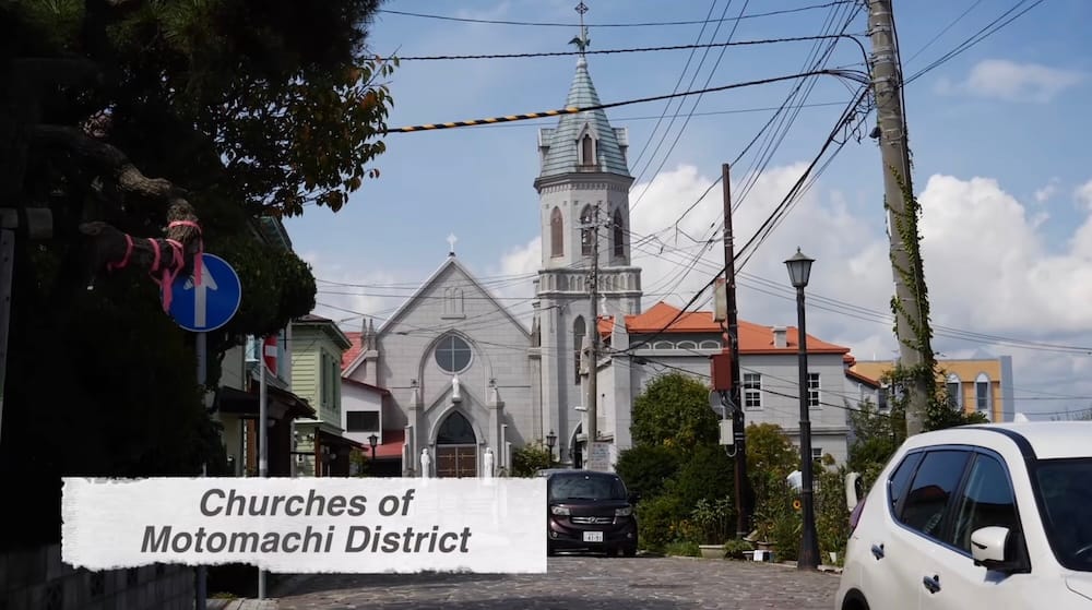 Motomachi Roman Catholic Church in Hakodate, Hokkaido, Japan, viewed along a quiet street with its pale Gothic facade, tall bell tower, red roof details, utility lines and surrounding buildings.