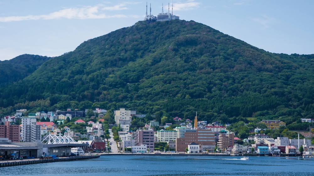 Mt. Hakodate Above the Historic Waterfront, Hokkaido, Japan – Forested Slopes, Summit Facilities and Harbour Mt. Hakodate rises above the waterfront in Hakodate, Hokkaido, Japan, with densely forested slopes, summit observation facilities and antennas overlooking colourful hillside buildings, harbour piers and calm blue water.