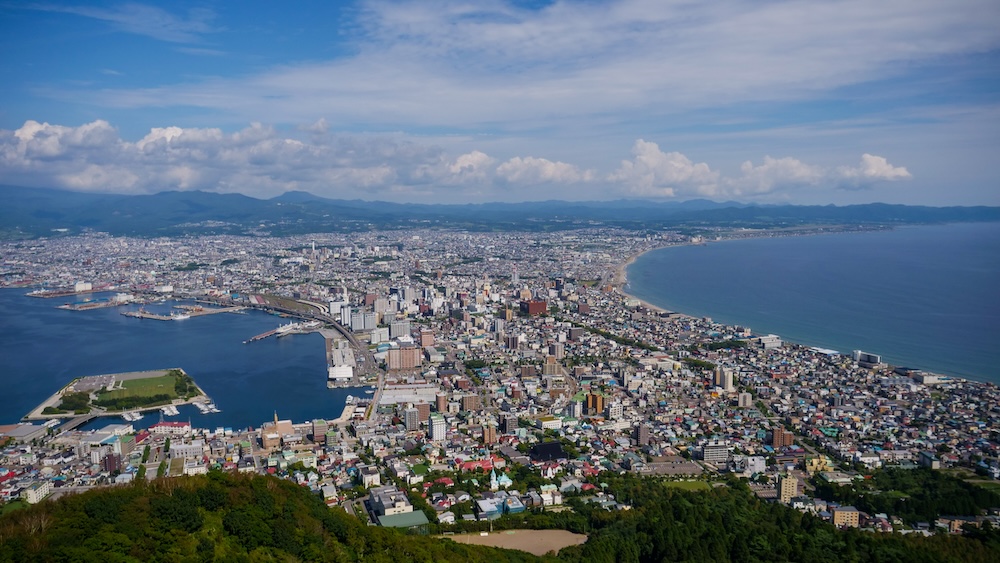 Panoramic View From Mt. Hakodate, Hokkaido, Japan – City Between Hakodate Harbour and the Tsugaru Strait Panoramic daytime view from Mt. Hakodate in Hokkaido, Japan, showing the densely built city stretching inland between Hakodate Harbour on the left and the sweeping coastline of the Tsugaru Strait on the right, with distant mountains.