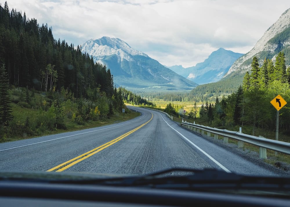 View through a car windshield of an empty two-lane mountain highway curving toward snow-streaked peaks and dense forest in the Canadian Rockies near Banff National Park, Alberta, Canada.