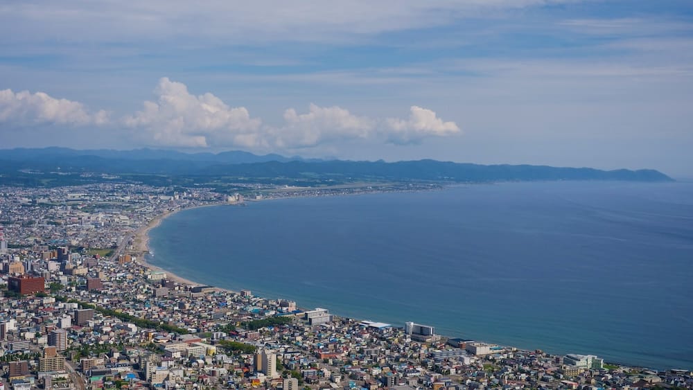 Mt. Hakodate daytime view over Hakodate coastline, city, bay and distant mountains in Hokkaido, Japan Daytime view from Mt. Hakodate in Hakodate, Hokkaido, Japan, showing dense city neighbourhoods curving along the coastline beside a broad blue bay, with distant mountains beneath scattered clouds.