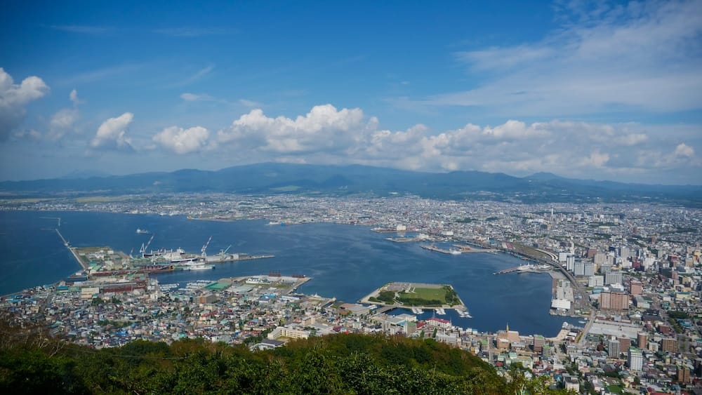 Daytime panorama from Mt. Hakodate over Hakodate harbour, port and city with distant mountains in Hokkaido, Japan Daytime panorama from Mt. Hakodate in Hakodate, Hokkaido, Japan, overlooking the harbour, port facilities, dense city neighbourhoods and distant mountains beneath a blue, cloud-filled sky.