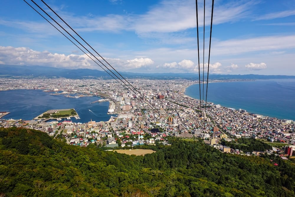 Ropeway cables run down the forested slope of Mt. Hakodate toward the harbour, coastline and dense city in Hakodate, Hokkaido, Japan, with mountains on the horizon and scattered clouds in a blue sky.