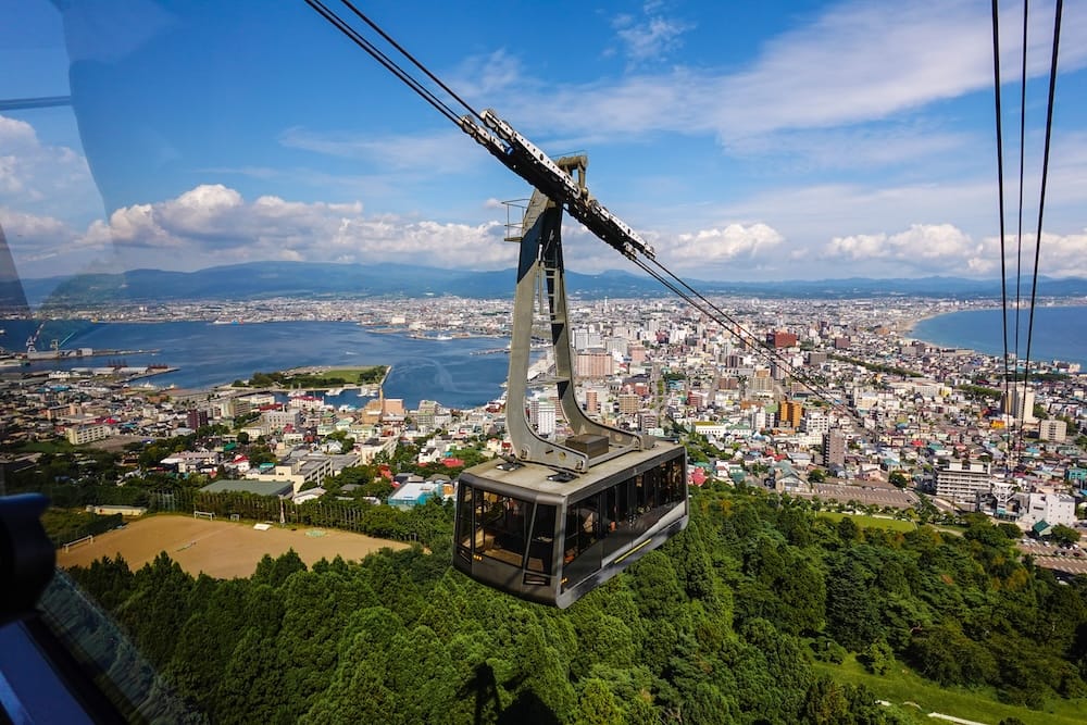 A Mt. Hakodate Ropeway gondola passes over forested slopes in Hakodate, Hokkaido, Japan, with the city, harbour, a stretch of coastline and distant mountains spread out beneath a blue sky with scattered clouds.