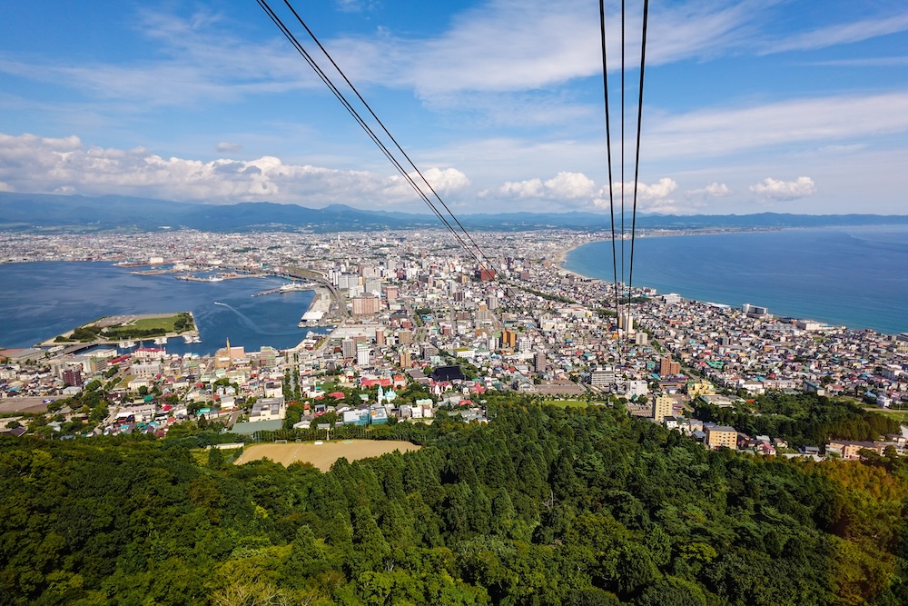 Panoramic daytime view from the Mt. Hakodate Ropeway in Hakodate, Hokkaido, Japan, showing cable lines above forested slopes, dense city neighbourhoods, harbour waters and the sweeping coastline on both sides.