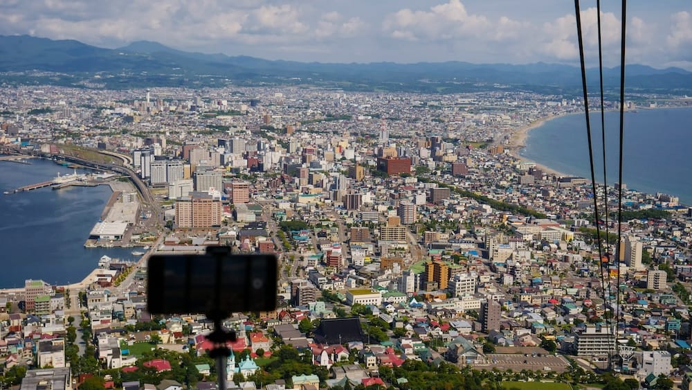 View from the Mt. Hakodate Ropeway above Hakodate, Hokkaido, Japan, showing the city, harbour and curving coastline below, with ropeway cables and a silhouetted camera or phone mount in the foreground.