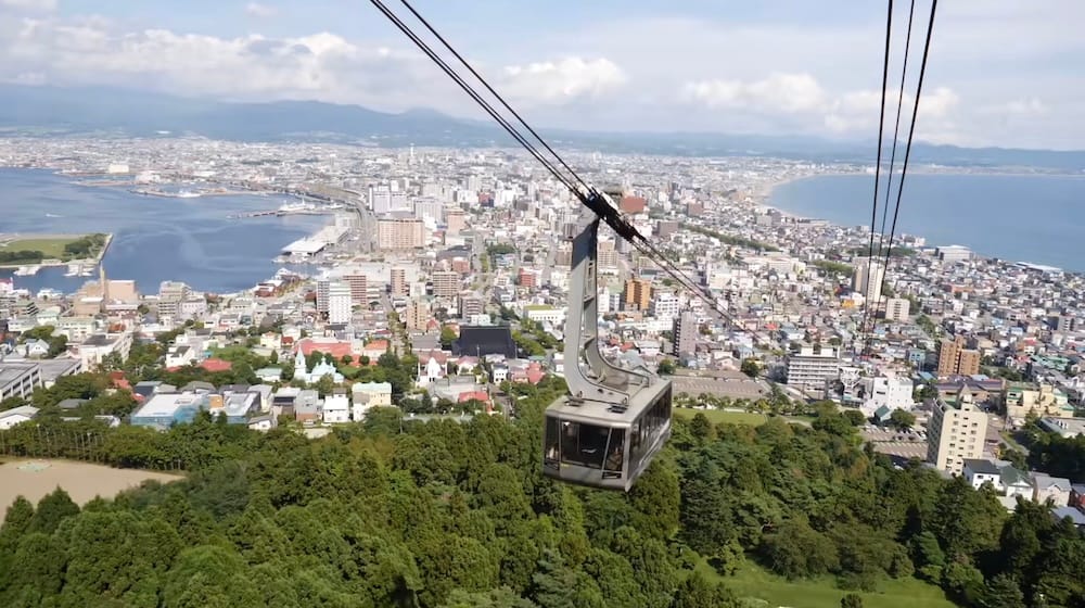 Mt. Hakodate Ropeway gondola suspended above Hakodate, Hokkaido, Japan, with the city spread between the harbour and curving coastline below and mountains visible beyond under partly cloudy skies.