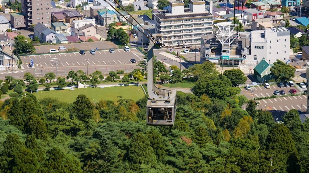 A Mt. Hakodate Ropeway gondola descends above the green hillside toward Hakodate, Hokkaido, Japan, with residential streets, parking areas and city buildings visible below.