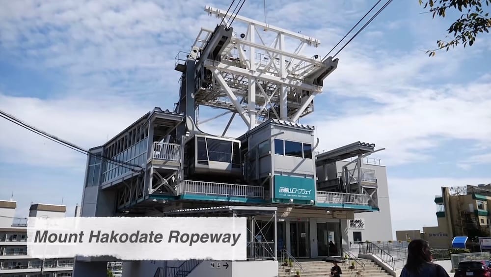 Exterior of the Mt. Hakodate Ropeway station in Hakodate, Hokkaido, Japan, with a gondola at the terminal, overhead cables, metal support structure, stairs and the station building under a partly cloudy sky.
