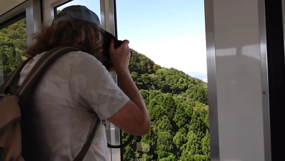 Samuel Jeffery photographs the view through the window while riding the Mt. Hakodate Ropeway in Hakodate, Hokkaido, Japan, with dense green forest covering the hillside below.