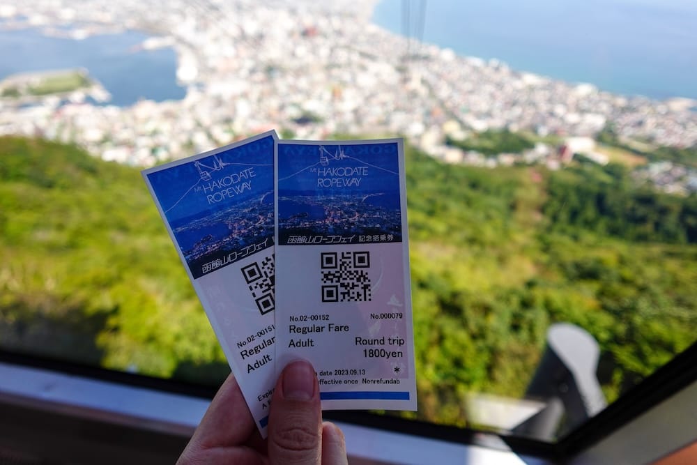 Two Mt. Hakodate Ropeway round-trip tickets are held inside the gondola during the ascent above Hakodate, Hokkaido, Japan, with the green mountainside and a blurred city-and-bay panorama visible through the window.