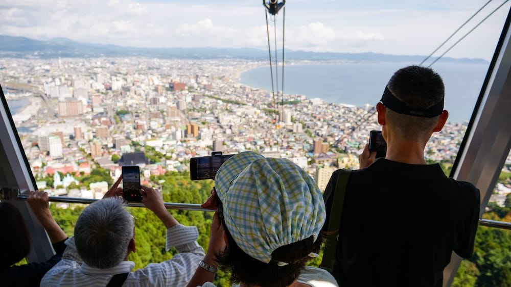 Tourists photographing Hakodate city and coastline from the Mt. Hakodate Ropeway in Hokkaido, Japan Tourists photograph the daytime view from the Mt. Hakodate Ropeway in Hakodate, Hokkaido, Japan, with the city, curved coastline, harbour and distant mountains visible through the windows.