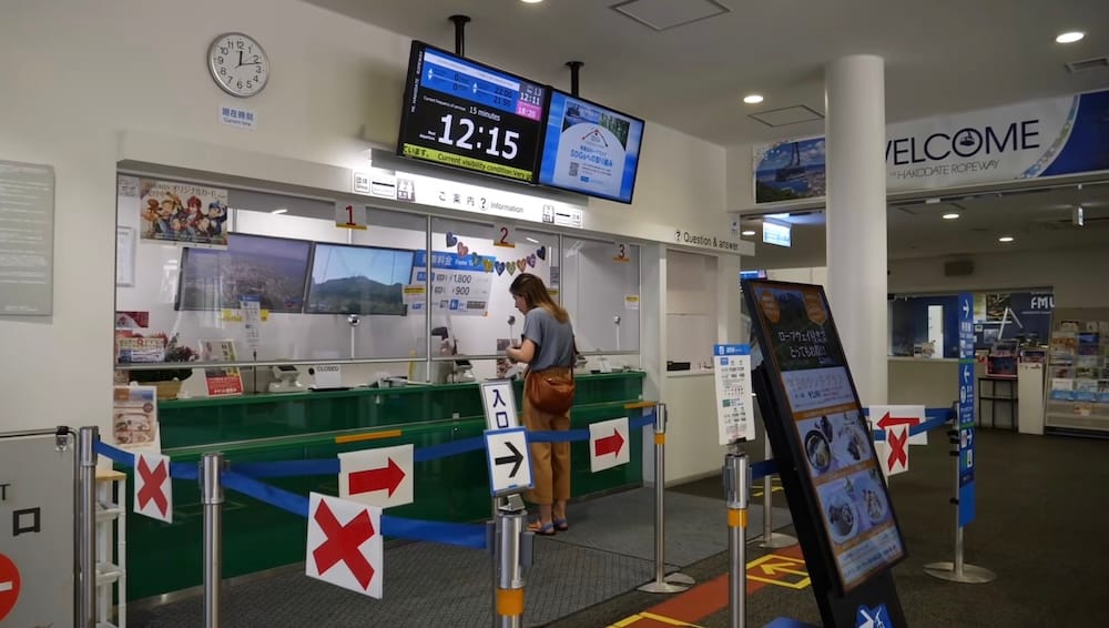 Audrey Bergner comprando entradas para el Mt. Hakodate Ropeway en Hakodate, Hokkaido, Japón Audrey Bergner comprando entradas en la boletería del Mt. Hakodate Ropeway en Hakodate, Hokkaido, Japón, rodeada de carteles de tarifas, pantallas informativas e indicaciones de embarque.