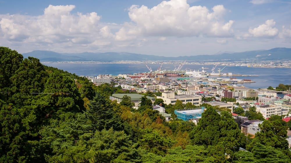 Vista diurna en altura sobre el paisaje urbano de Hakodate, grúas del puerto y la bahía desde el monte Hakodate, Hokkaido, Japón Vista diurna desde el monte Hakodate mirando sobre laderas boscosas hacia la ciudad de Hakodate, el puerto con grúas y montañas lejanas en Hokkaido, Japón, bajo un cielo azul con nubes blancas.