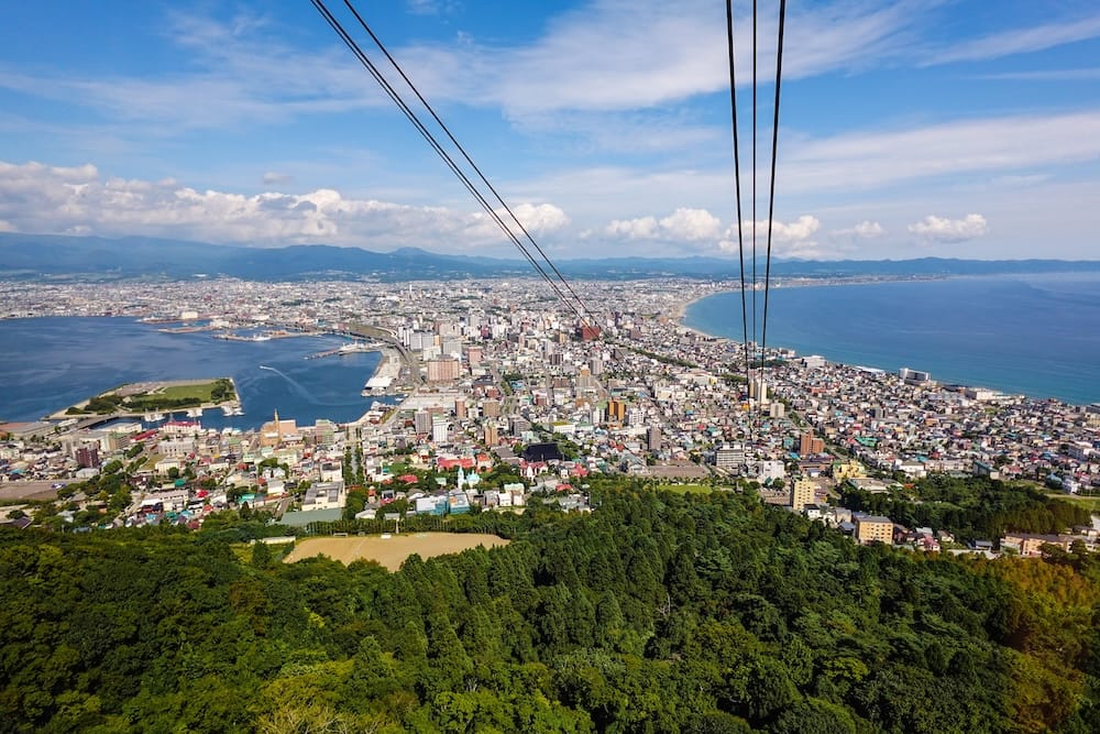 Ciudad, puerto y lado de la playa vistos desde la góndola del teleférico del monte Hakodate, Hakodate, Hokkaido, Japón La vista desde una góndola del teleférico del monte Hakodate sobre Hakodate, Hokkaido, Japón, con los cables en primer plano, la ladera boscosa abajo, la ciudad, el puerto a la izquierda, la curva de la playa a la derecha y montañas de fondo.