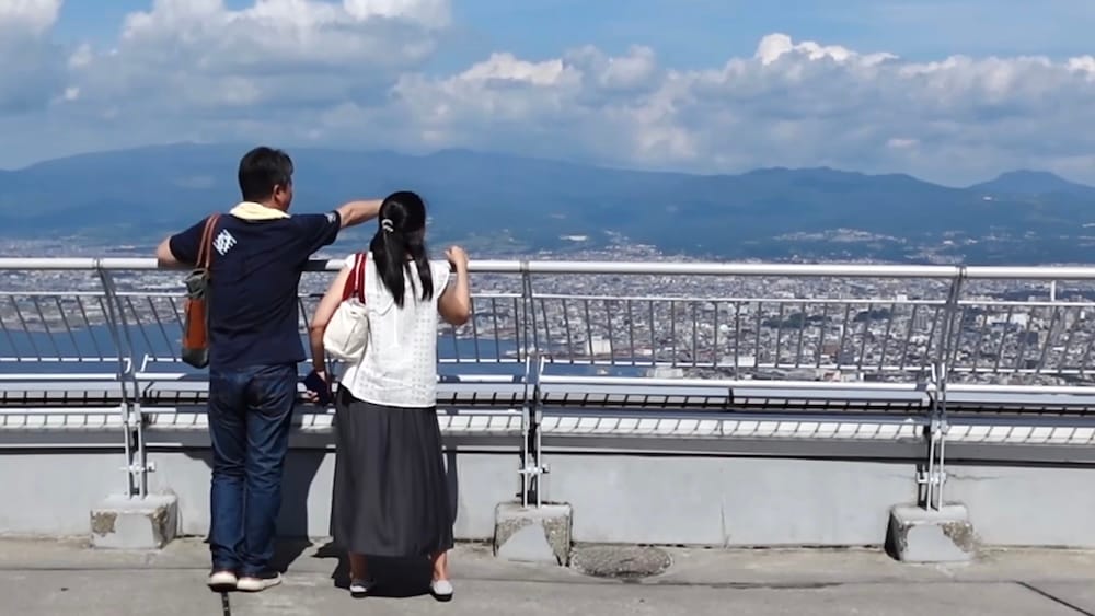 Visitantes en la baranda del mirador de la cima del monte Hakodate, Hakodate, Hokkaido, Japón Dos visitantes se apoyan en la baranda del mirador de la cima del monte Hakodate en Hakodate, Hokkaido, Japón, mirando hacia la ciudad y las montañas bajo un cielo azul con nubes grandes.