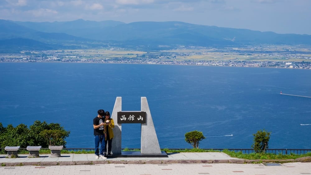 Daytime view from the Mt. Hakodate viewpoint in Hakodate, Hokkaido, Japan, with two visitors beside the summit sign and the blue bay, distant shoreline and mountain range stretching across the background.