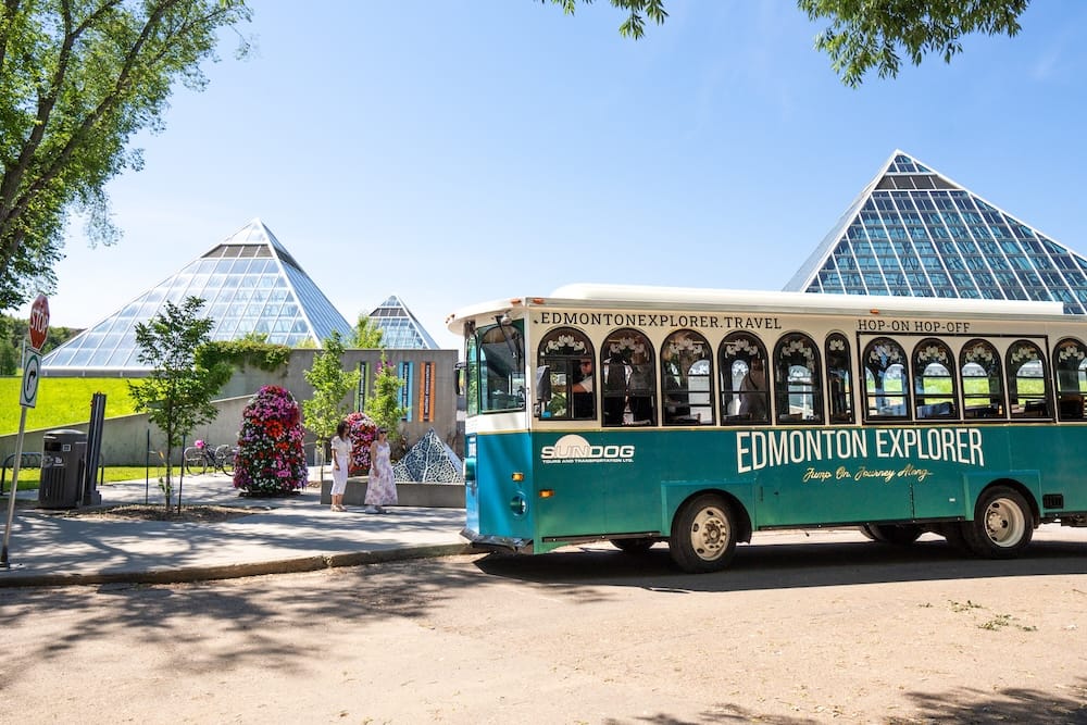 Edmonton Explorer trolley at Muttart Conservatory in Edmonton, Alberta, Canada Edmonton Explorer hop-on hop-off trolley parked beside the glass pyramids of the Muttart Conservatory in Edmonton, Alberta, Canada, with trees, pedestrians and summer flowers nearby.