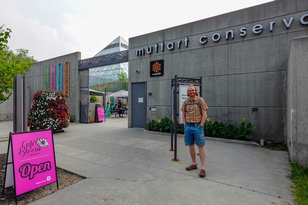 Samuel Jeffery standing outside the entrance to Muttart Conservatory in Edmonton, Alberta, Canada, with the concrete façade, glass pyramid, flower display and café sign visible around him.