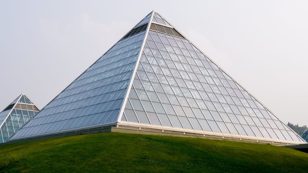 Exterior view of the glass pyramids at Muttart Conservatory in Edmonton, Alberta, Canada, rising above a green grassy slope with geometric panes reflecting the pale sky.