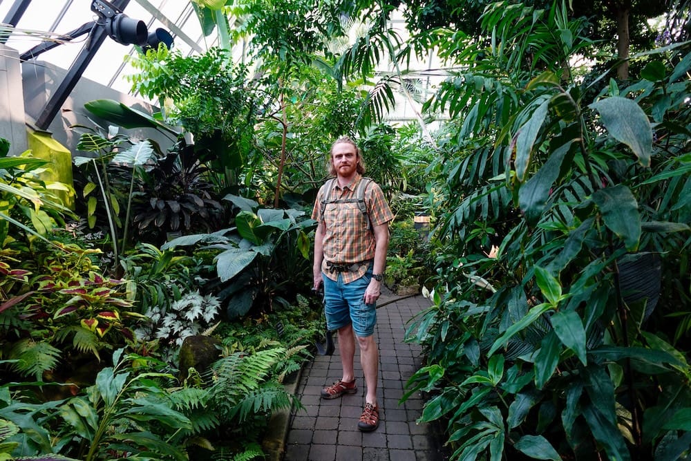 Samuel Jeffery stands on a brick path inside the Tropical Pyramid at Muttart Conservatory in Edmonton, Alberta, Canada, surrounded by dense green foliage, broad-leaf plants and the glass greenhouse structure overhead.