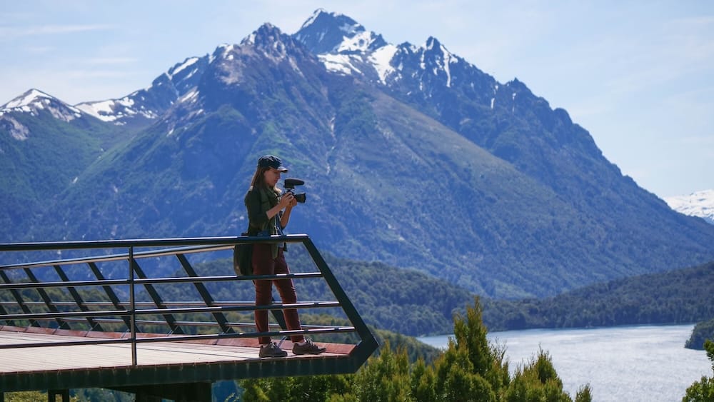 Audrey Bergner filming Nahuel Huapi Lake and mountain views in Bariloche, Río Negro, Patagonia, Argentina Audrey Bergner films from a viewing platform above Nahuel Huapi Lake, with forested slopes and snow-streaked mountains rising behind her in Bariloche, Río Negro, Patagonia, Argentina.