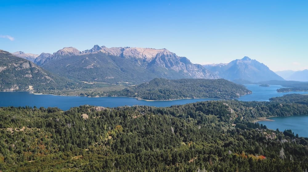 Panoramic Nahuel Huapi Lake and Andean peaks near Bariloche, Patagonia, Argentina A panoramic view across Nahuel Huapi Lake near Bariloche, Río Negro, Patagonia, Argentina, shows deep blue water winding between forested peninsulas and broad Andean ridges beneath a clear sky.