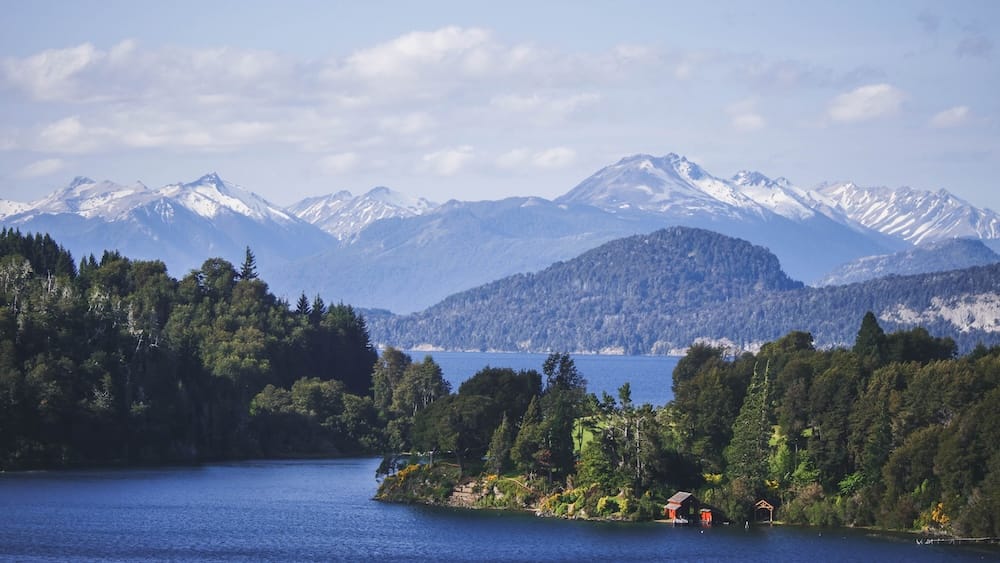 Una casita roja para botes aparece entre un bosque denso junto al lago Nahuel Huapi en San Carlos de Bariloche, Río Negro, Patagonia, Argentina, con picos andinos y restos de nieve al otro lado del agua.