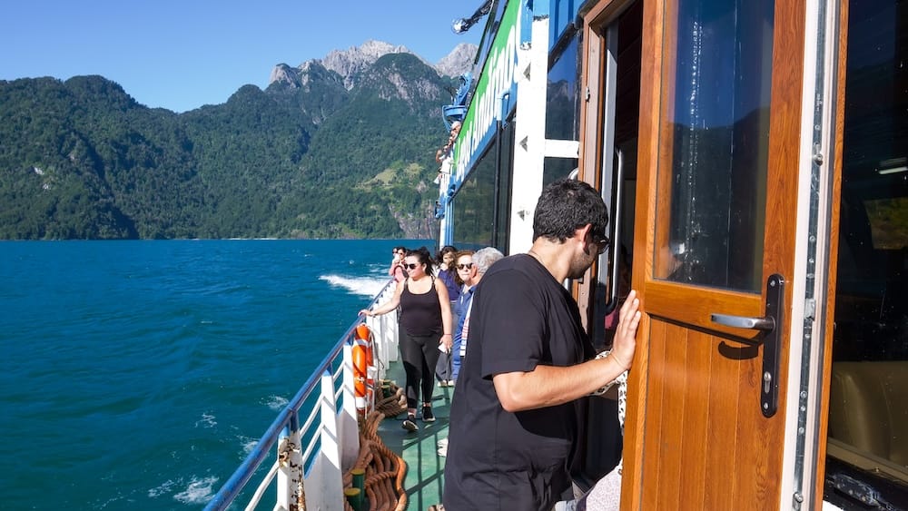 Passengers stand on the deck of a boat crossing Nahuel Huapi Lake near Bariloche, Río Negro, Patagonia, Argentina, with vivid turquoise water and densely forested mountain slopes rising behind them.