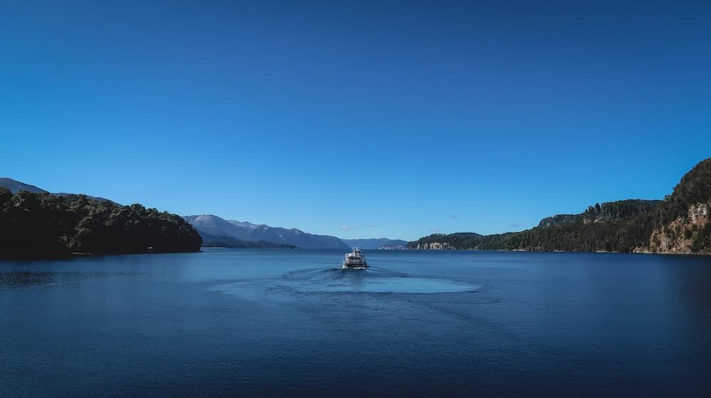A catamaran crosses Nahuel Huapi Lake near Villa La Angostura, Neuquén, Patagonia, Argentina, leaving a wake across deep blue water between forested shores and distant mountain ridges.