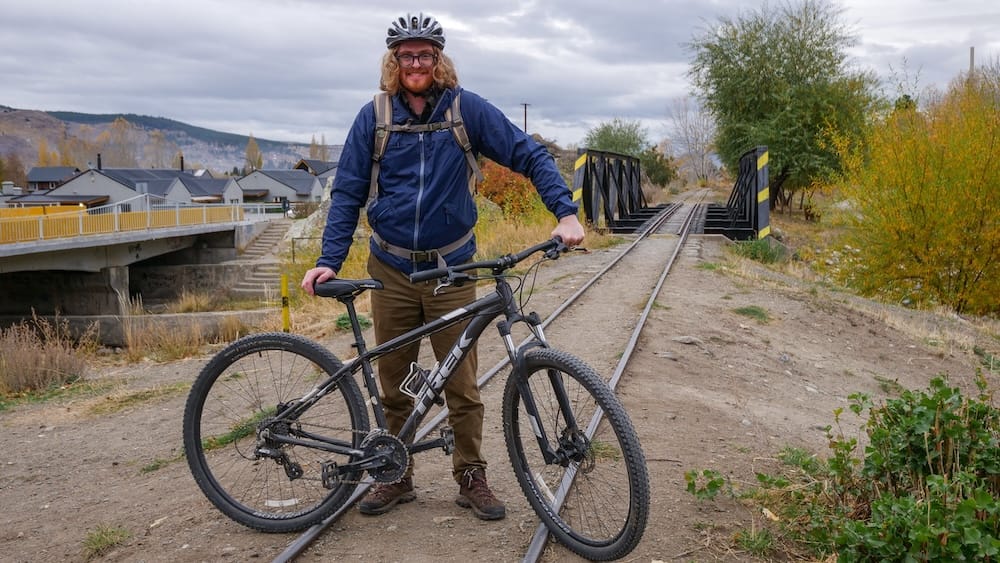 Samuel Jeffery stands with a mountain bike on narrow-gauge railway tracks in Esquel, Chubut, Patagonia, Argentina, wearing a helmet and backpack beneath grey skies beside a small rail bridge and autumn trees.