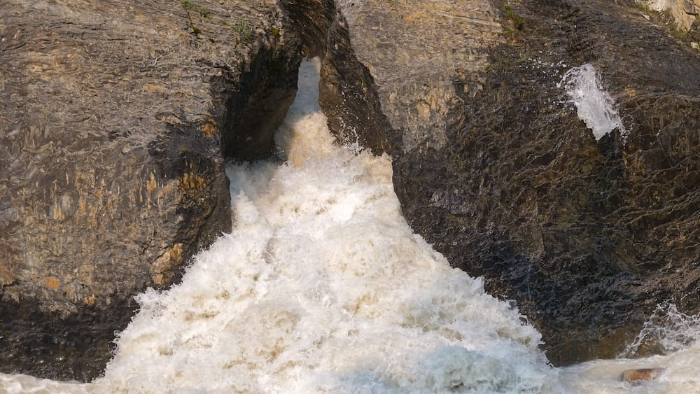 Close view of the Kicking Horse River surging through a narrow rock channel at Natural Bridge in Yoho National Park, British Columbia, Canada, with white water churning between dark exposed rock.